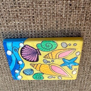 May include: A rectangular, hand-painted wooden block featuring a beach scene. The design includes a blue wave, seashells, a starfish, and sand. The seashells are pink, orange, and blue, with a yellow background.