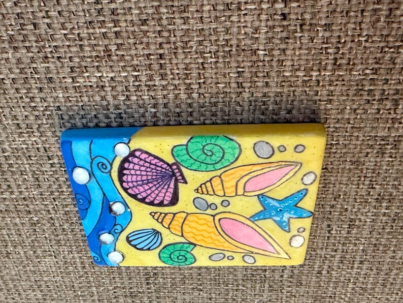 May include: A rectangular, hand-painted wooden block featuring a beach scene. The design includes a blue wave, seashells, a starfish, and sand. The seashells are pink, orange, and blue, with a yellow background.