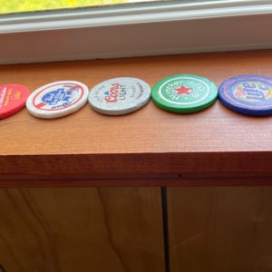 Puede incluir: Cinco tapas redondas de botellas de cerveza de diferentes colores con diferentes nombres de marca. Las tapas est&aacute;n dispuestas en fila sobre una superficie de madera. Las marcas son Budweiser, Pabst Blue Ribbon, Coors Light, Heineken y Miller Lite.