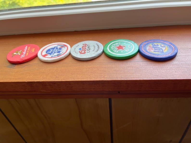 Puede incluir: Cinco tapas redondas de botellas de cerveza de diferentes colores con diferentes nombres de marca. Las tapas est&aacute;n dispuestas en fila sobre una superficie de madera. Las marcas son Budweiser, Pabst Blue Ribbon, Coors Light, Heineken y Miller Lite.