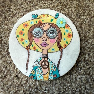 Puede incluir: Posavasos redondo y blanco con una ilustraci&oacute;n de una mujer con un sombrero floral amarillo y gafas. La mujer tiene trenzas marrones y una chaqueta turquesa con detalles florales. Un colgante con el s&iacute;mbolo de la paz cuelga de su cuello.