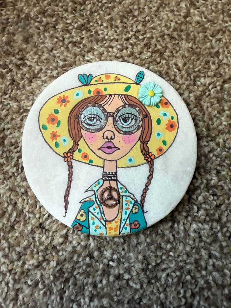 Puede incluir: Posavasos redondo y blanco con una ilustraci&oacute;n de una mujer con un sombrero floral amarillo y gafas. La mujer tiene trenzas marrones y una chaqueta turquesa con detalles florales. Un colgante con el s&iacute;mbolo de la paz cuelga de su cuello.