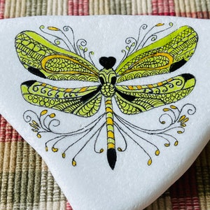 K&ouml;nnte beinhalten: Ein handbemalter Stein mit einem detaillierten gr&uuml;nen und gelben Libellen-Design. Die Libelle hat schwarze Akzente und komplizierte Muster auf ihren Fl&uuml;geln.