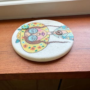 Puede incluir: Un posavasos redondo y blanco con una colorida ilustraci&oacute;n de una mujer con un sombrero amarillo con flores y gafas azules. La mujer tiene trenzas largas y un elemento decorativo en su camisa. El posavasos est&aacute; sobre una superficie de madera.