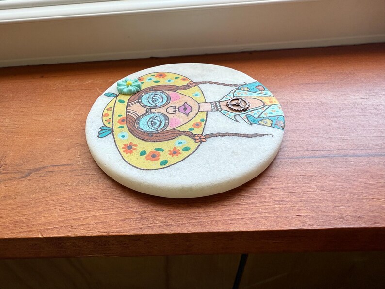 Puede incluir: Un posavasos redondo y blanco con una colorida ilustraci&oacute;n de una mujer con un sombrero amarillo con flores y gafas azules. La mujer tiene trenzas largas y un elemento decorativo en su camisa. El posavasos est&aacute; sobre una superficie de madera.