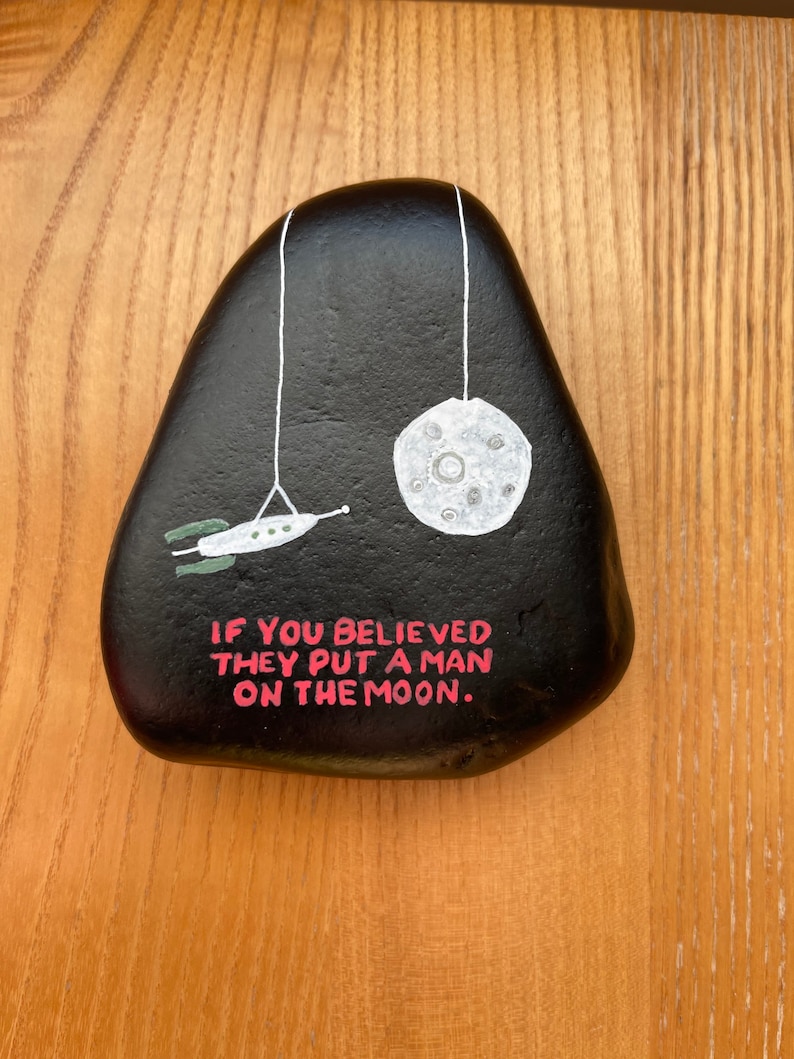 May include: A black painted rock with a white moon and a rocket ship painted on it. The rock has the text "If you believed they put a man on the moon." written on it in pink.