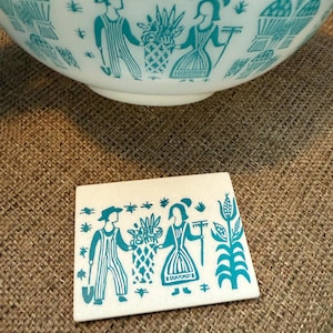 以下が含まれることがあります： 白いボウルと、花かごを持つカップルのターコイズ色のイラストが描かれたお揃いのコースター。ボウルは同じターコイズのデザインの広い縁があります。コースターは長方形です。