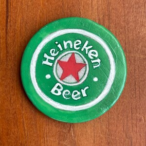 Puede incluir: Un posavasos redondo verde Heineken con un c&iacute;rculo blanco alrededor del borde. El c&iacute;rculo tiene la palabra "Heineken" escrita en letras blancas y la palabra "Beer" escrita en letras blancas debajo de una estrella roja.