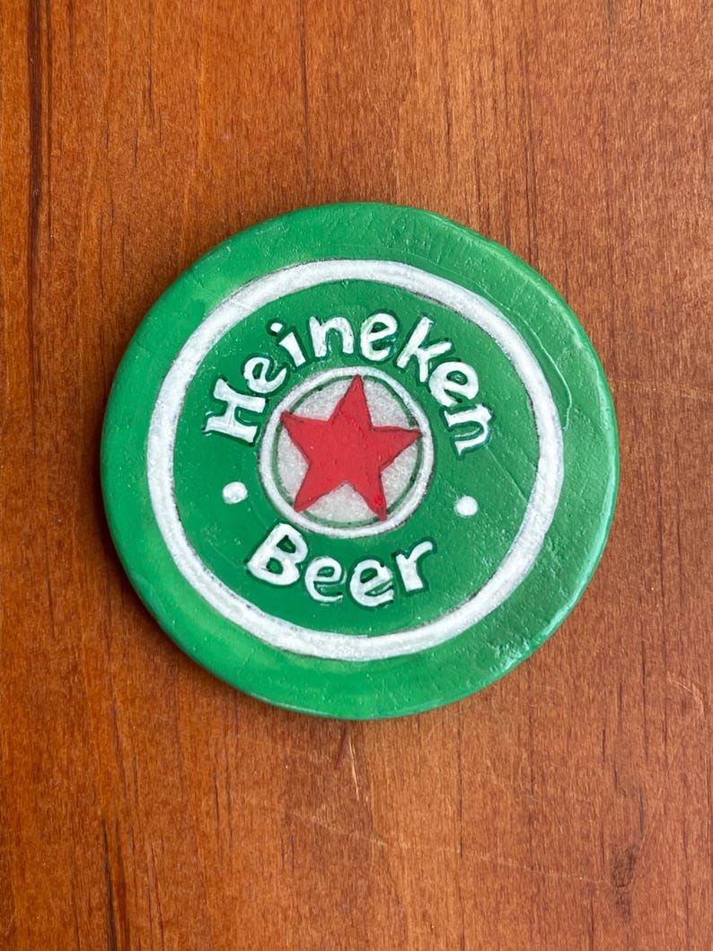 Puede incluir: Un posavasos redondo verde Heineken con un c&iacute;rculo blanco alrededor del borde. El c&iacute;rculo tiene la palabra "Heineken" escrita en letras blancas y la palabra "Beer" escrita en letras blancas debajo de una estrella roja.