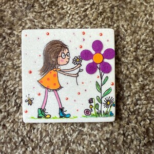 Puede incluir: Azulejo de cer&aacute;mica cuadrado con una ilustraci&oacute;n caprichosa de una ni&ntilde;a con gafas con un vestido naranja, recogiendo una flor. Una gran flor morada con un centro naranja est&aacute; a su lado. Tambi&eacute;n se representa una abeja y otras flores.