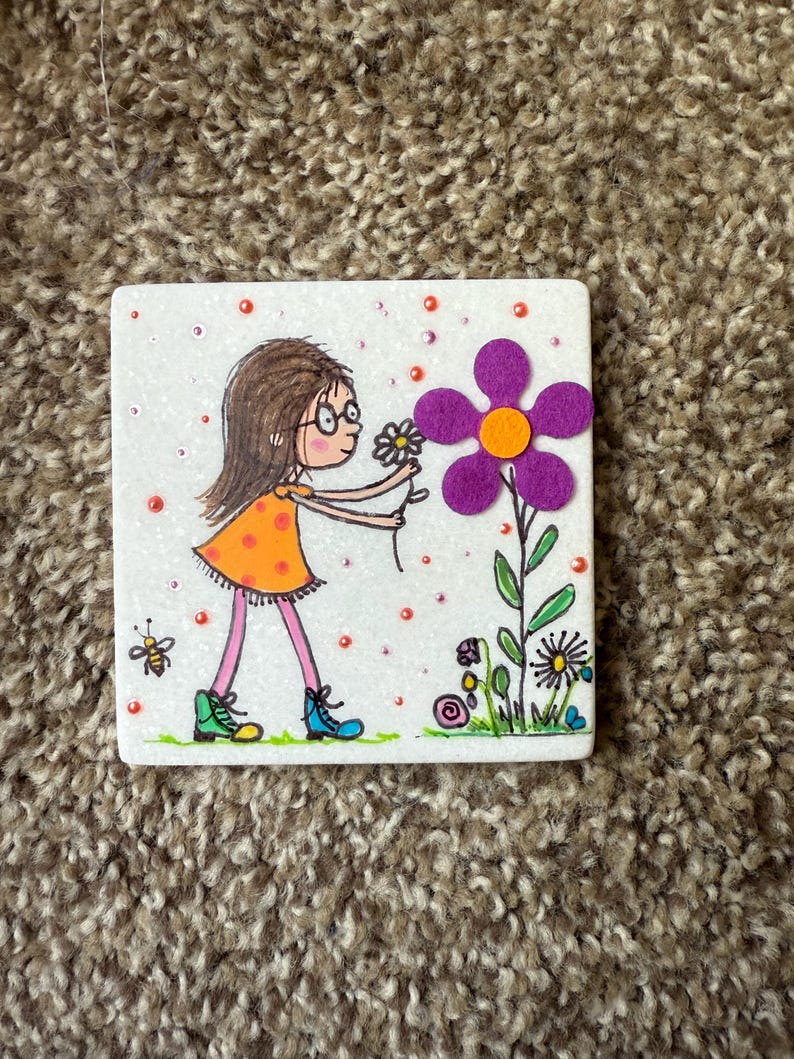 Puede incluir: Azulejo de cer&aacute;mica cuadrado con una ilustraci&oacute;n caprichosa de una ni&ntilde;a con gafas con un vestido naranja, recogiendo una flor. Una gran flor morada con un centro naranja est&aacute; a su lado. Tambi&eacute;n se representa una abeja y otras flores.