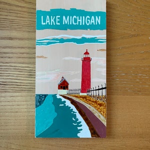 K&ouml;nnte beinhalten: Ein Holzschild mit dem Text "LAKE MICHIGAN" in wei&szlig; auf einem t&uuml;rkisfarbenen Hintergrund. Ein roter Leuchtturm mit wei&szlig;em Dach und ein kleineres rotes Geb&auml;ude mit wei&szlig;em Dach sind auf das Schild gemalt. Die Geb&auml;ude befinden sich auf einem braunen Steg mit einer t&uuml;rkisfarbenen Welle, die dagegen schl&auml;gt.