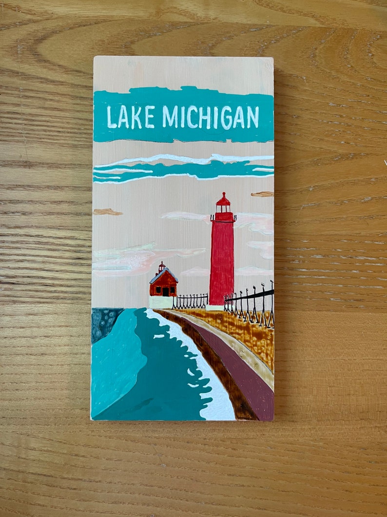 K&ouml;nnte beinhalten: Ein Holzschild mit dem Text "LAKE MICHIGAN" in wei&szlig; auf einem t&uuml;rkisfarbenen Hintergrund. Ein roter Leuchtturm mit wei&szlig;em Dach und ein kleineres rotes Geb&auml;ude mit wei&szlig;em Dach sind auf das Schild gemalt. Die Geb&auml;ude befinden sich auf einem braunen Steg mit einer t&uuml;rkisfarbenen Welle, die dagegen schl&auml;gt.