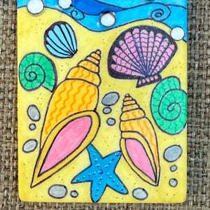 May include: A rectangular, hand-painted tile featuring a beach scene. The design includes yellow sand, blue waves, and various seashells in orange, pink, green, and blue. A blue starfish and small gray stones complete the artwork.