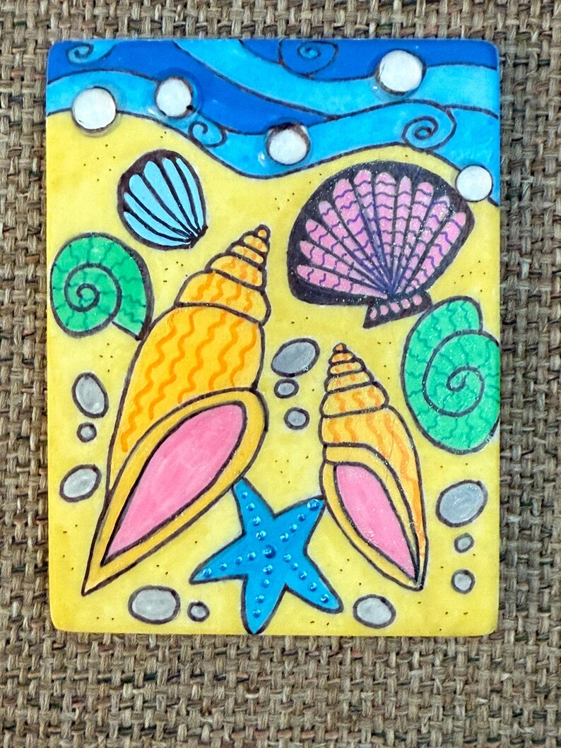 May include: A rectangular, hand-painted tile featuring a beach scene. The design includes yellow sand, blue waves, and various seashells in orange, pink, green, and blue. A blue starfish and small gray stones complete the artwork.
