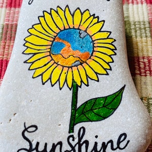 Puede incluir: Piedra pintada con un girasol amarillo con un centro azul y naranja. El texto "You are my sunshine" est&aacute; escrito en negro debajo de la flor.