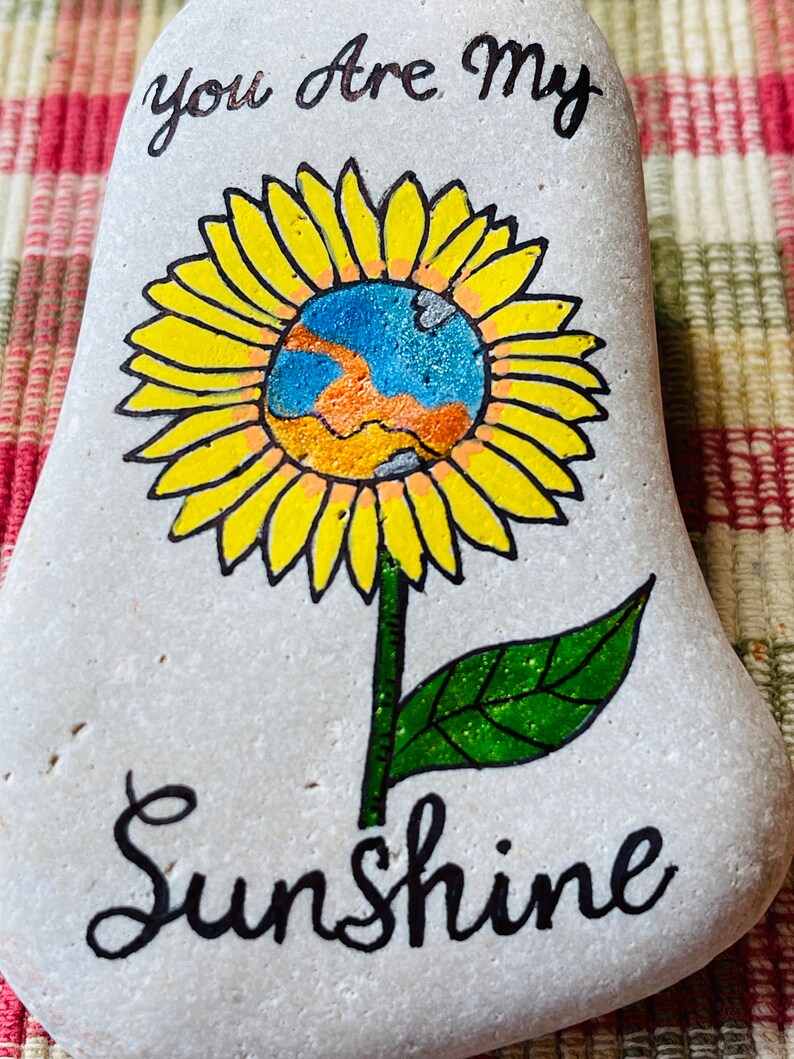 Puede incluir: Piedra pintada con un girasol amarillo con un centro azul y naranja. El texto "You are my sunshine" est&aacute; escrito en negro debajo de la flor.