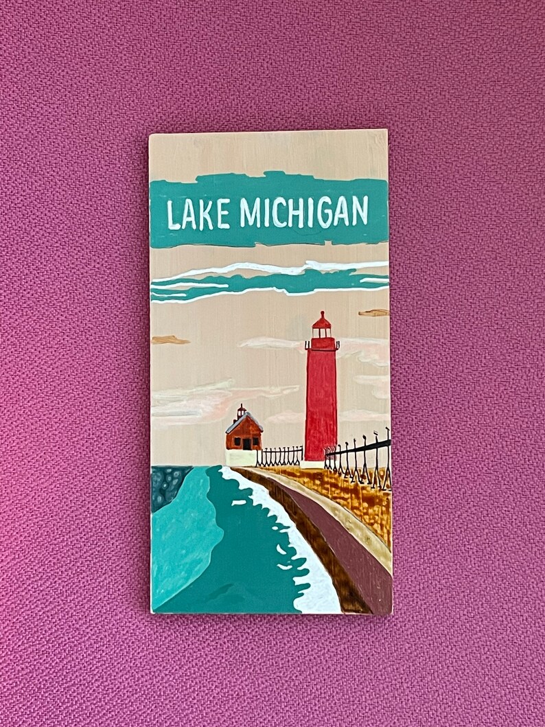 K&ouml;nnte beinhalten: Ein Holzschild mit dem Text "LAKE MICHIGAN" in wei&szlig; auf einem t&uuml;rkisfarbenen Hintergrund. Das Schild zeigt einen roten Leuchtturm mit einem wei&szlig;en Dach und ein kleineres wei&szlig;es Geb&auml;ude mit einem roten Dach. Der Leuchtturm steht auf einem Steg mit einem braunen Holzgel&auml;nder. Das Wasser ist t&uuml;rkisfarben mit wei&szlig;en Wellen.