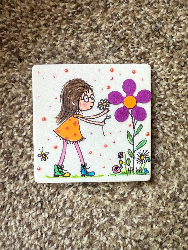 Puede incluir: Azulejo de cer&aacute;mica cuadrado con una ilustraci&oacute;n de dibujos animados de una ni&ntilde;a con un vestido naranja y botas azules recogiendo una flor. Tambi&eacute;n se representa una abeja y otras flores.