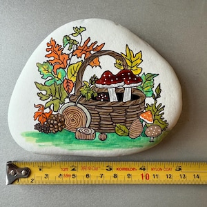 Puede incluir: Una piedra blanca pintada a mano presenta una escena oto&ntilde;al detallada. Una cesta tejida contiene setas de sombrero rojo con puntos blancos. Hojas coloridas, una pi&ntilde;a y rodajas de madera rodean la cesta. La piedra mide aproximadamente 13 cm de ancho.