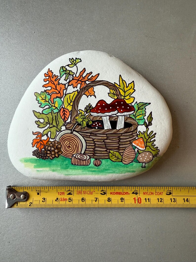 Puede incluir: Una piedra blanca pintada a mano presenta una escena oto&ntilde;al detallada. Una cesta tejida contiene setas de sombrero rojo con puntos blancos. Hojas coloridas, una pi&ntilde;a y rodajas de madera rodean la cesta. La piedra mide aproximadamente 13 cm de ancho.