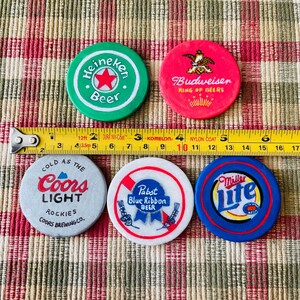 Puede incluir: Cinco fichas de cerveza redondas y de colores con diferentes logotipos. Las fichas son verde, roja, gris, blanca y azul. Los logotipos incluyen Heineken Beer, Budweiser King of Beers, Coors Light, Pabst Blue Ribbon Beer y Miller Lite.