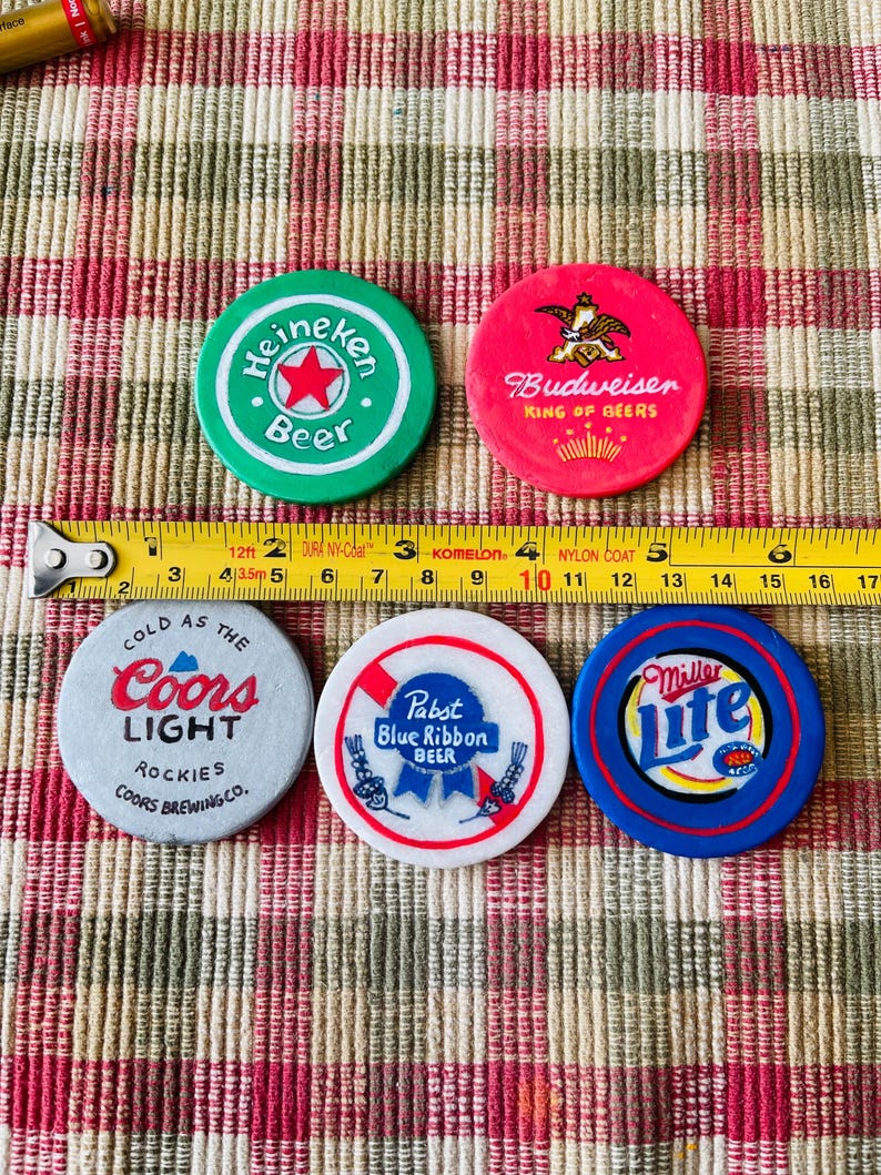 Puede incluir: Cinco fichas de cerveza redondas y de colores con diferentes logotipos. Las fichas son verde, roja, gris, blanca y azul. Los logotipos incluyen Heineken Beer, Budweiser King of Beers, Coors Light, Pabst Blue Ribbon Beer y Miller Lite.
