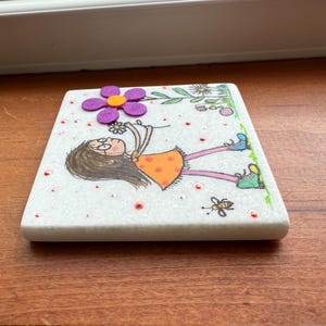Puede incluir: Un posavasos cuadrado de m&aacute;rmol blanco con una ilustraci&oacute;n caprichosa. Presenta una ni&ntilde;a de dibujos animados con un vestido naranja, sosteniendo una flor, con una flor morada y una abeja. El posavasos est&aacute; decorado con puntos rojos y detalles verdes.