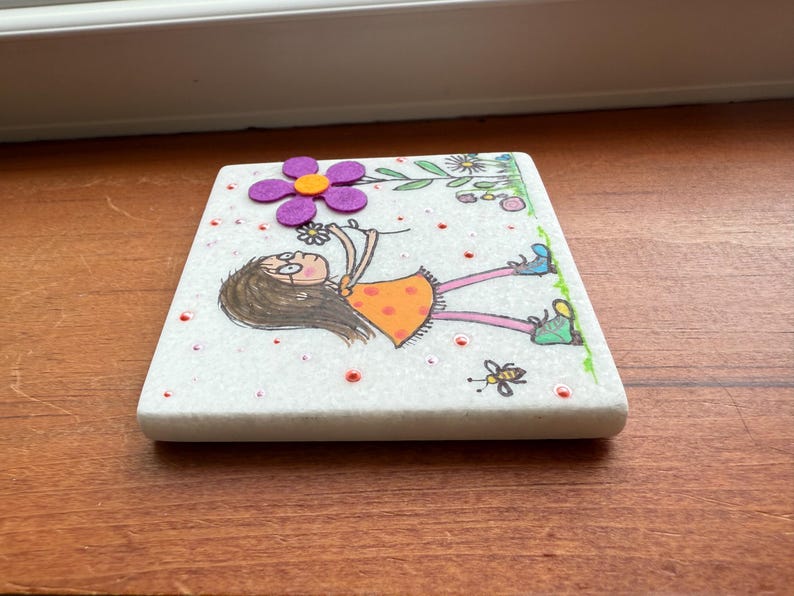 Puede incluir: Un posavasos cuadrado de m&aacute;rmol blanco con una ilustraci&oacute;n caprichosa. Presenta una ni&ntilde;a de dibujos animados con un vestido naranja, sosteniendo una flor, con una flor morada y una abeja. El posavasos est&aacute; decorado con puntos rojos y detalles verdes.