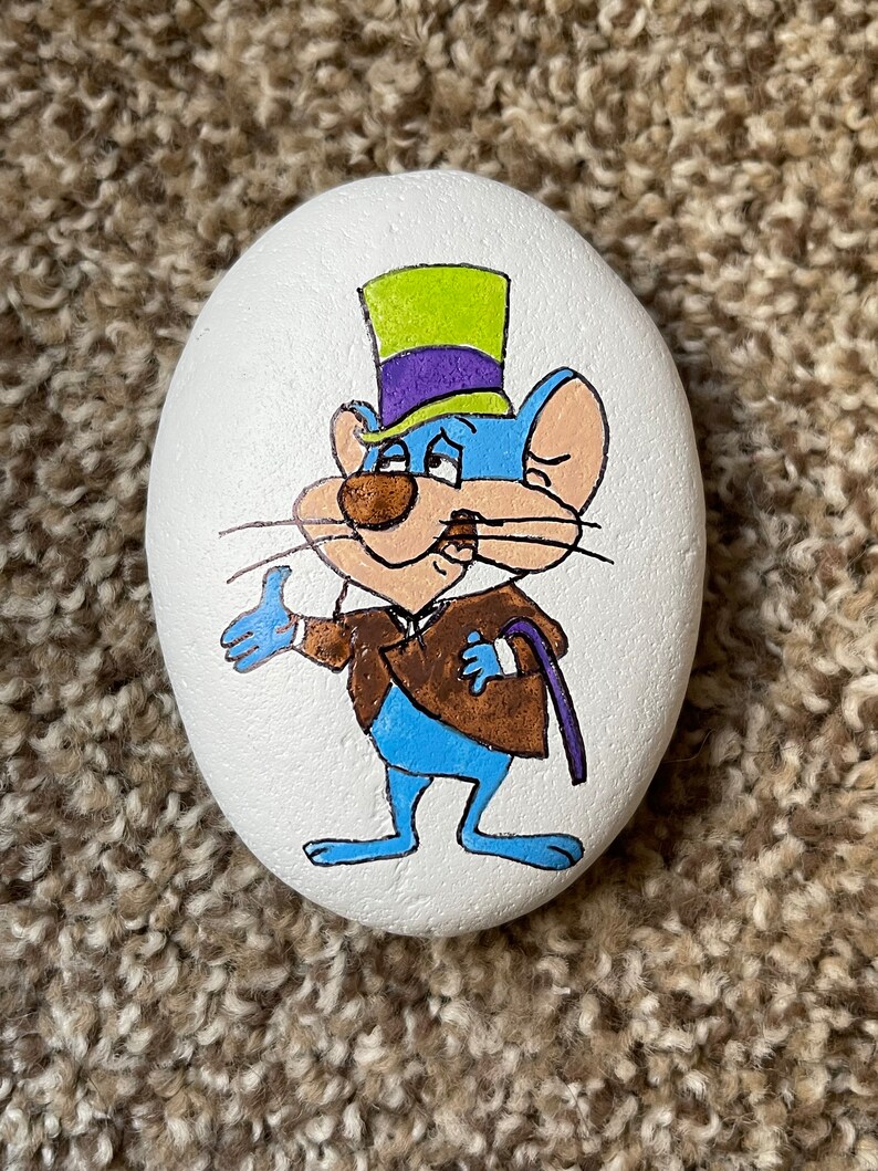 Puede incluir: Piedra pintada a mano con un rat&oacute;n de dibujos animados que lleva un sombrero de copa morado y verde, una chaqueta marr&oacute;n y pantalones azules. El rat&oacute;n sostiene un bast&oacute;n en su mano derecha y tiene una expresi&oacute;n de sorpresa en su rostro.