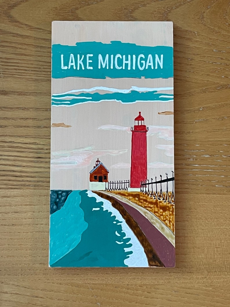 K&ouml;nnte beinhalten: Ein Holzschild mit einer gemalten Szene eines roten Leuchtturms auf einem Pier mit einem blauen See und wei&szlig;en Wolken. Das Schild tr&auml;gt die Aufschrift "LAKE MICHIGAN" in wei&szlig;en Buchstaben.