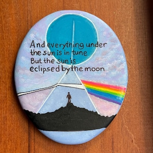 May include: A round, hand-painted stone featuring a blue moon, a black silhouette of a person on a hill, and a rainbow prism effect. The text reads: And everything under the sun is in tune. But the sun is eclipsed by the moon. The background is light purple.