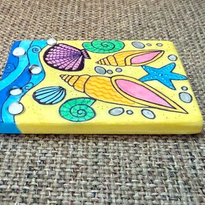 May include: A rectangular, hand-painted wooden plaque with a beach theme. The design features yellow sand, blue waves, and various seashells, a starfish, and spiral shells in vibrant colors. The edges are painted blue.