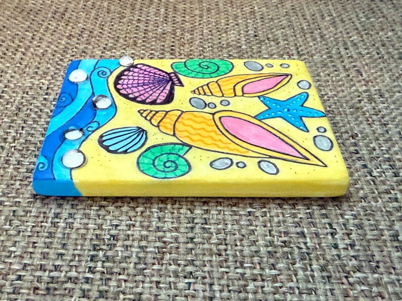 May include: A rectangular, hand-painted wooden plaque with a beach theme. The design features yellow sand, blue waves, and various seashells, a starfish, and spiral shells in vibrant colors. The edges are painted blue.