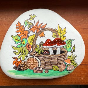Puede incluir: Una piedra blanca pintada a mano con un dise&ntilde;o de oto&ntilde;o. Una cesta tejida contiene dos setas con sombrero rojo y puntos blancos. Hojas coloridas y otras setas rodean la cesta. Una pieza decorativa para el hogar.