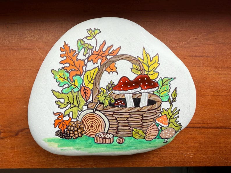 Puede incluir: Una piedra blanca pintada a mano con un dise&ntilde;o de oto&ntilde;o. Una cesta tejida contiene dos setas con sombrero rojo y puntos blancos. Hojas coloridas y otras setas rodean la cesta. Una pieza decorativa para el hogar.