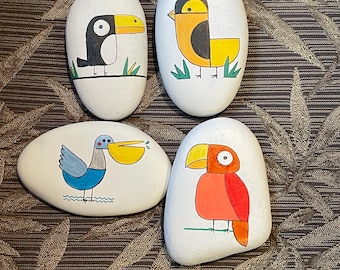 May include: Four painted rocks with different bird designs. The birds are a toucan, a yellow bird, a blue bird with a yellow beak, and a red bird with a yellow beak. The rocks are all white and have a smooth surface.