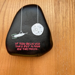 May include: A black painted rock with a white moon and a spaceship hanging from it. The rock has the text "If you believed they put a man on the moon." written on it in pink.