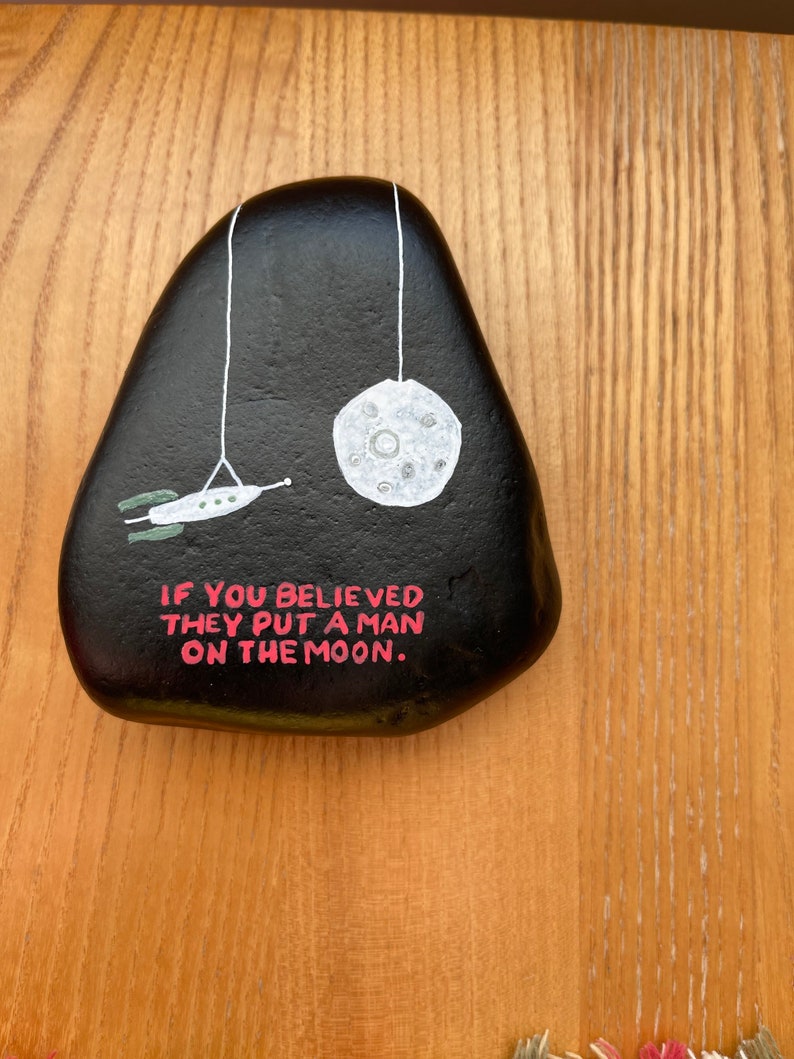 May include: A black painted rock with a white moon and a spaceship hanging from it. The rock has the text "If you believed they put a man on the moon." written on it in pink.