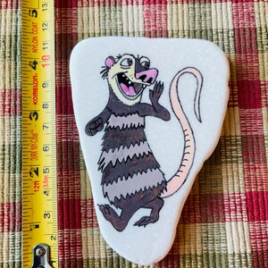 May include: Hand-painted rock art featuring a cartoon opossum with a pink nose and tail. The opossum is depicted with a surprised expression and is standing on its hind legs. The rock is white with a smooth surface.