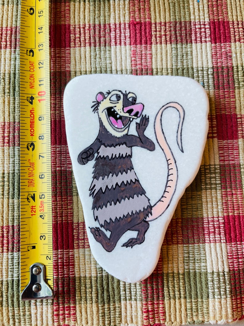 May include: Hand-painted rock art featuring a cartoon opossum with a pink nose and tail. The opossum is depicted with a surprised expression and is standing on its hind legs. The rock is white with a smooth surface.