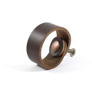 May include: A brown leather cabinet knob with a copper screw. The knob is shaped like a ring with a small copper button in the center.