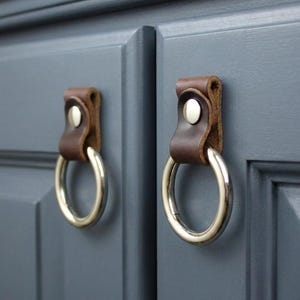 May include: Two cabinet pulls with brown leather straps and silver-colored ring handles. The pulls are attached to a gray cabinet door. The leather straps are secured with silver-colored rivets.