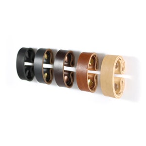 May include: Five leather strap holders in different colors: black, dark brown, light brown, tan, and cream. Each holder has two brass rivets.