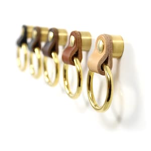 May include: Five brass key hooks with leather straps in different shades of brown. The hooks are mounted on a white wall.
