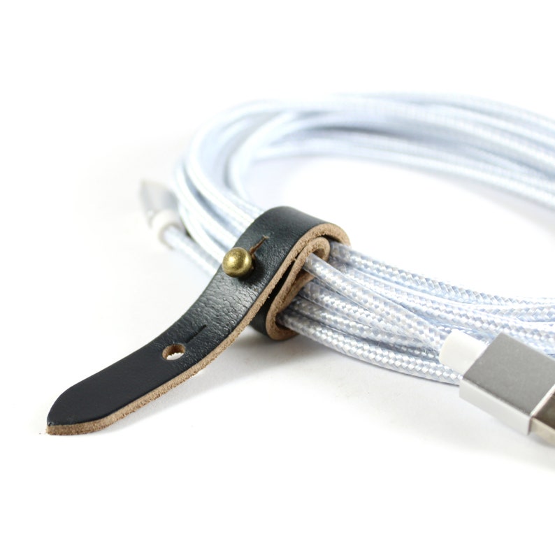 May include: A white braided cable with a black leather strap and a brass button. The cable is coiled and the strap is wrapped around it.
