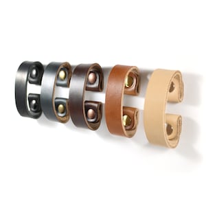 May include: Five leather strap handles in different colors, including black, gray, brown, tan, and beige. Each handle has two brass rivets and a C-shape design.
