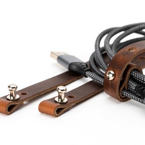 May include: Two brown leather straps with metal snaps, used to organize and secure a gray braided cable. The cable has a gray and silver connector.