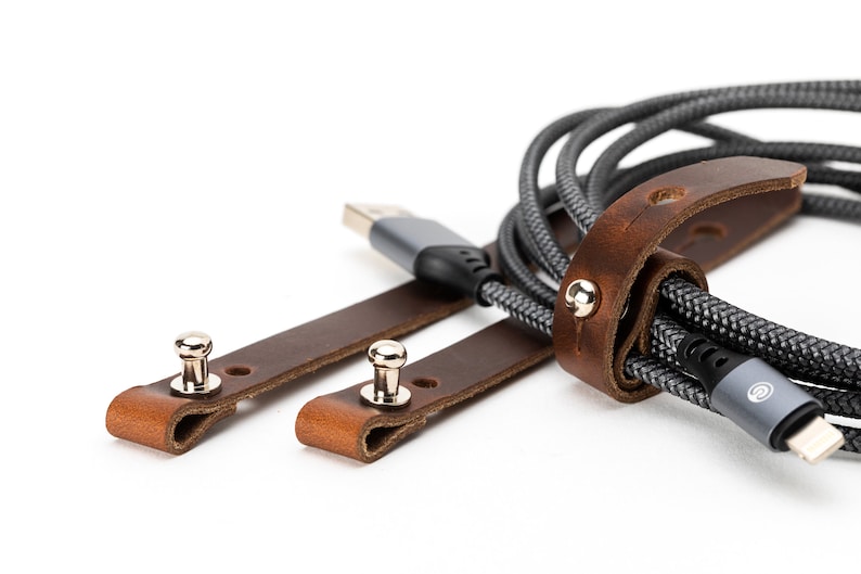 May include: Two brown leather straps with metal snaps, used to organize and secure a gray braided cable. The cable has a gray and silver connector.