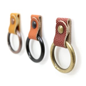 May include: Three leather key rings with different colored leather straps and metal rings. The rings are black, brass, and copper.